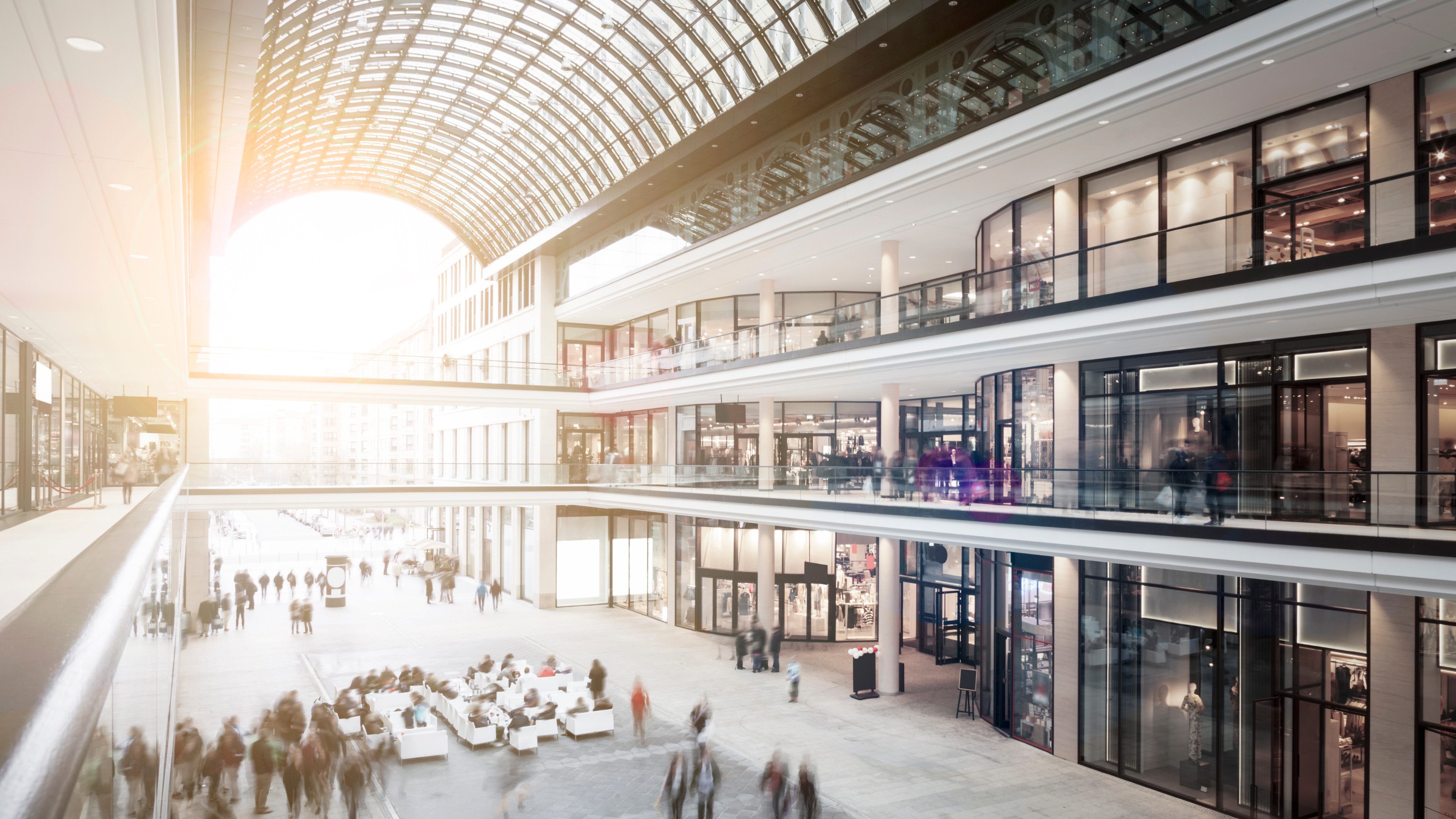 Modern shopping mall with outdoor seating and three floors of stores. There are motioned blurred people sitting, shopping and walking around. The architecture is modern and elegant.